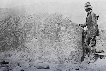 Un hombre con sombrero y ropa de expedición toca una gran roca cubierta de petroglifos. La imagen en blanco y negro muestra un paisaje montañoso y árido de fondo