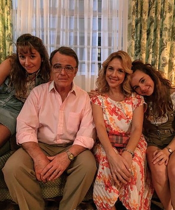 Un hombre mayor con gafas y camisa rosa posa sentado en un sillón junto a tres mujeres en un entorno hogareño