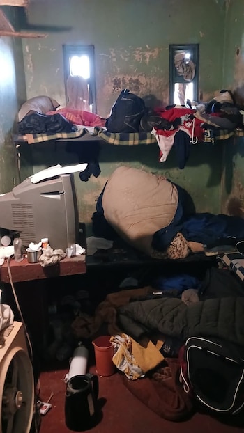 Habitación con literas cubiertas de ropa y equipaje, un televisor antiguo sobre un estante y pertenencias dispersas en el suelo