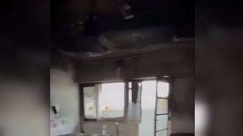 Interior de habitación con techo y paredes oscurecidas por el humo y daños estructurales, ventanas visibles y objetos en el suelo