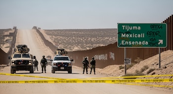 Vehículos militares y policiales con luces encendidas, junto a personal uniformado, en un camino de terracería en una zona fronteriza árida, con cinta policial y un letrero de carretera con grafiti.