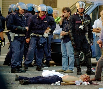Hombre en el suelo con heridas visibles, rodeado por policías con cascos azules y un carabinero con máscara de gas. Un fotógrafo y un vehículo policial están cerca