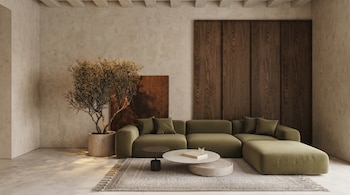 Una sala de estar con paredes beige y suelo de cemento, sofá seccional verde oliva, mesa de centro redonda, planta alta en maceta y panel de madera oscuro.