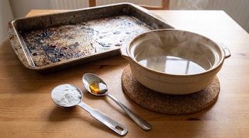 Cuchara con bicarbonato, cuchara con jabón líquido, cazuela con agua caliente y bandeja con grasa quemada sobre una mesa de madera.