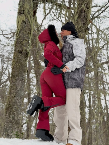 Un hombre y una mujer con ropa de invierno posan abrazados sobre la nieve en un bosque