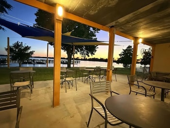 Patio exterior con mesas y sillas. Fondo de un lago, muelle y árboles al atardecer, con cielo naranja y azul. Luces de la pérgola encendidas