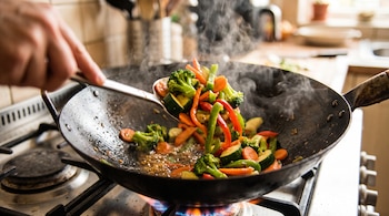 Un wok metálico sobre un fogón encendido contiene brócoli, zanahoria, pimiento y calabacín mientras una mano los remueve con una espátula.