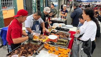 Dos hombres con gorros y delantales preparan chicharrón en un puesto callejero. Múltiples bandejas con chicharrón, camotes fritos y tamales. Clientes esperan