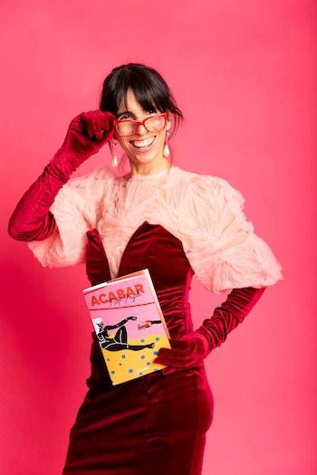 Una mujer con gafas rojas, guantes largos de terciopelo y vestido sostiene un libro titulado Acabar frente a un fondo rosa