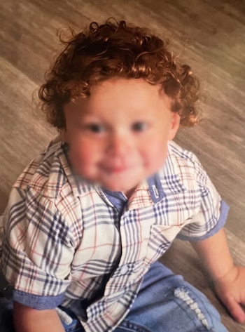 Un niño de cabello rizado viste una camisa a cuadros clara y un pantalón azul mientras se apoya sentado sobre una superficie de madera