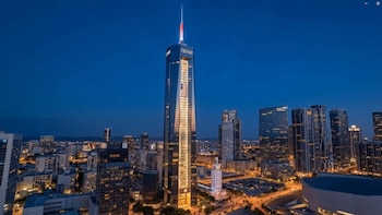 Vista aérea nocturna de un rascacielos moderno con el nombre 'TRUMP' iluminado y una bandera estadounidense en su fachada, rodeado de edificios urbanos