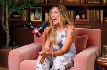 Karol G, con cabello rubio y top de lentejuelas, sentada en un sillón rosa riendo con la boca abierta frente a un micrófono de podcast