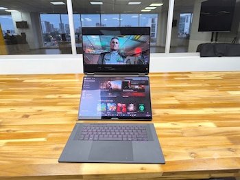 Un portátil gris ASUS ROG Zephyrus Duo abierto sobre una mesa de madera, con una pantalla principal mostrando un videojuego y una pantalla secundaria con interfaz de juegos