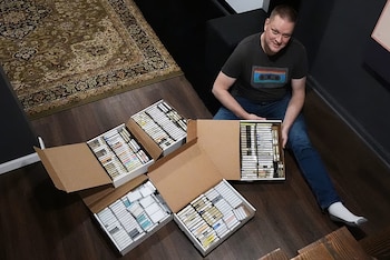 Hombre sentado en el suelo de madera, sonriendo, rodeado de cuatro cajas de cartón llenas de cintas de casete. Lleva una camiseta con diseño de casete