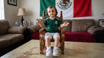 Estatua del Niño Dios sentada en silla de madera. Viste camiseta verde de la selección mexicana, shorts blancos y tenis. Detrás, bandera de México.