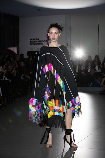 Una modelo camina por una pasarela con una prenda negra decorada con flecos multicolores ante la mirada del público