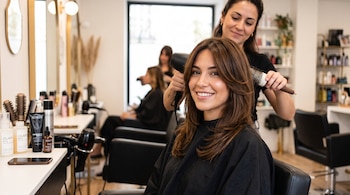 Una peluquera seca con secador y cepillo redondo el cabello castaño de una mujer sentada en un salón con espejos y productos.