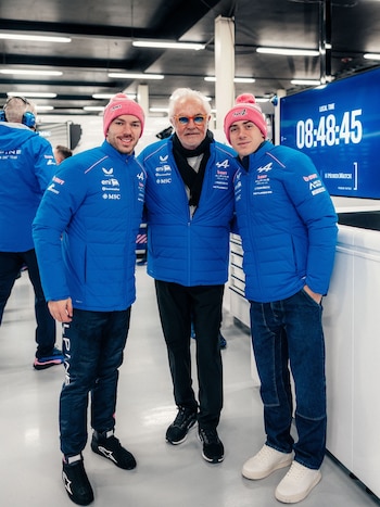 Tres hombres, dos jóvenes con chaquetas azules y gorros rosados, uno mayor con chaqueta azul y gafas de sol, posan en un pasillo junto a una pantalla digital