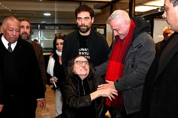 Charly García, sentado en silla de ruedas, sonríe con gafas y chaqueta de cuero. Pedro Aznar, de pie con bufanda roja, toca su brazo. Hay otras personas
