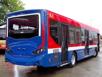 Un autobús moderno azul y rojo de la marca Ugarte, con múltiples puertas, estacionado sobre un suelo de cemento mojado. Se observa parte de otro autobús en el fondo