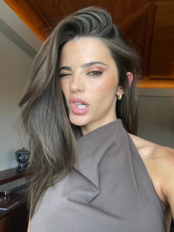 Inés Aguiar, mujer de cabello castaño largo y oscuro, guiña un ojo derecho y entreabre los labios. Viste un top gris sin mangas y un aro dorado. Fondo borroso