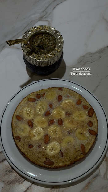 Vista aérea de una torta de avena con rodajas de plátano y almendras en un plato blanco, junto a un mate tradicional con bombilla sobre una superficie de mármol