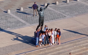 Vista aérea de la estatua de bronce de Rocky Balboa con los brazos levantados, rodeada por un grupo de adultos y niños posando en una plaza pavimentada