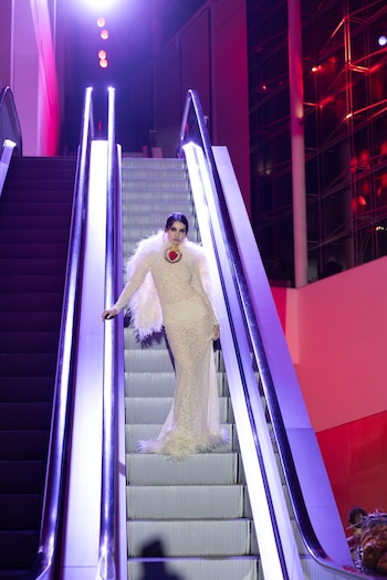 Una mujer vestida con un traje largo claro de encaje y plumas posa de pie en el centro de una escalera mecánica iluminada por luces moradas