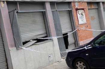 Persianas metálicas dañadas en la fachada de un local acordonado con cinta policial y un automóvil aparcado a la derecha