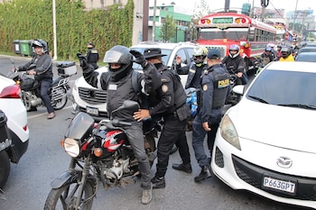 Varios agentes de la PNC inspeccionan a motociclistas en una calle con tráfico denso. Se observan coches blancos, un autobús rojo y vegetación verde
