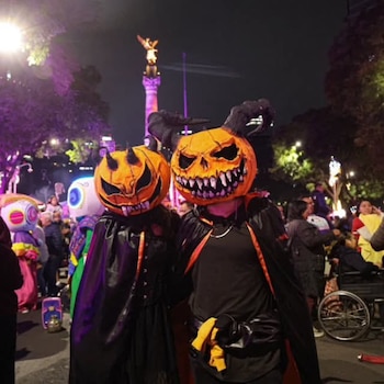 Dos personas con máscaras de calabaza y capas negras posan de noche frente al Ángel de la Independencia entre una multitud