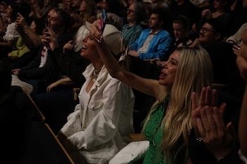 Dos mujeres rubias, una con chaqueta blanca y gorra beige y otra con chaqueta verde, ríen y graban con un teléfono en un evento lleno de público