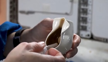 Dos manos manipulan un recipiente claro con líquido en su interior mediante un tubo transparente frente a paneles de instrumental blanco