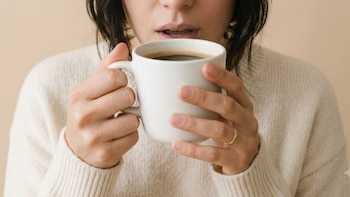 Boca y manos de una persona bebiendo café de una taza blanca. La persona viste un suéter de punto color crema y lleva un anillo dorado