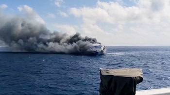 Ferry en el mar con fuego y humo negro. Cielo con nubes. Agua azul. Parte de una embarcación en primer plano a la derecha