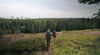 Dos personas con mochilas caminan por un sendero de tierra. Hay pasto seco, arbustos verdes y un denso bosque de coníferas al fondo bajo un cielo nublado