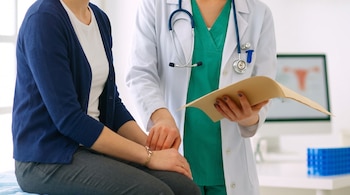 Una médica con bata y estetoscopio sostiene una carpeta y apoya su mano sobre la mano de una paciente en un consultorio.