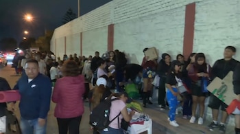 Varias personas con mochilas, colchonetas y cartones forman una fila nocturna junto a un muro blanco.