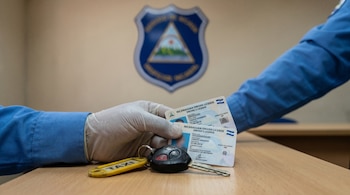 Mano enguantada de un agente sostiene dos licencias de conducir de Nicaragua y una llave de taxi en mesa de madera clara. Al fondo, logo desenfocado de la Policía Nacional de Nicaragua.