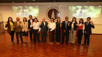 Un grupo de once personas alineadas en un escenario saluda con la mano levantada frente a dos pantallas de proyección