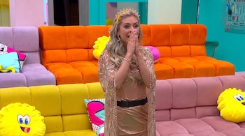 Una mujer rubia vestida de dorado y tiara se lleva las manos a la cara frente a sofás de colores amarillo, rosa y naranja.