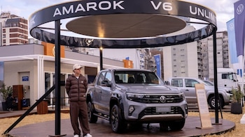 Pickup Volkswagen Amarok V6 gris en un stand de exhibición. Un joven con gorra y chaqueta de pie. Edificios, banderas Volkswagen y un camión blanco se ven al fondo
