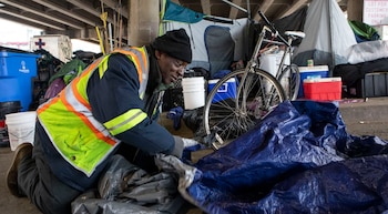 WM, un hombre con chaleco de alta visibilidad y gorro negro, se arrodilla en el suelo bajo un puente, organizando lonas azules y grises. Hay tiendas y una bicicleta detrás