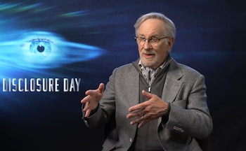 Steven Spielberg, un hombre mayor con gafas y barba, gesticula con las manos frente a un fondo oscuro con un ojo azul brillante y el texto "DISCLOSURE DAY"