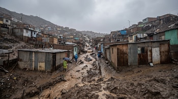 Asentamiento informal en ladera de cerro con casas de chapa y madera, caminos de lodo y agua. Varias personas con paraguas y ponchos transitan bajo cielo nublado