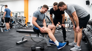 Tres hombres en un gimnasio. Uno está sentado en un banco de pesas sujetando su muslo con dolor. Dos hombres lo observan de pie. Pesas en el suelo.
