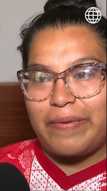 Primer plano de mujer con cabello oscuro recogido, gafas de montura clara y camiseta roja y blanca. Un micrófono es visible abajo a la izquierda