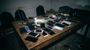 Mesa de madera oscura llena de numerosos teléfonos inteligentes en una habitación tenuemente iluminada, con una linterna que proyecta luz sobre ellos y mochilas al fondo.