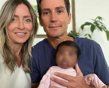 Primer plano de una mujer rubia sonriente junto a un hombre sonriente que sostiene a un bebé de piel oscura, con un fondo claro y una planta