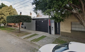 Frente de una casa con portón negro, pared gris y un árbol en la vereda junto a la calle